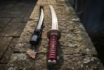 Hand forged high carbon steel katana with red wrapped handle and brass tsuba, angled view beside its black saya on stone