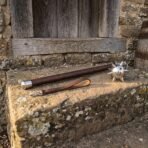 Hand forged high carbon steel medieval flail with walnut haft and leather wrist strap, angled view along a sunlit stone step
