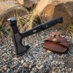 Hand forged high carbon steel tactical tomahawk standing head up on gravel, showing the etched head, spike and leather sheath