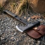 Hand forged high carbon steel tactical tomahawk with knotwork etched head, close view on gravel beside its leather sheath