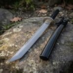 Hand forged high carbon steel tanto, close view of the angled tip on a mossy rock beside its black lacquered saya