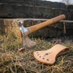 Hand forged high carbon steel tomahawk head with hammer poll, close view resting on dry grass with its laced tan leather cover