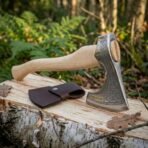 Hand forged high carbon steel Viking bearded axe with decorated head, close view resting on a birch log with its leather sheath