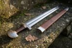 Hand forged high carbon steel Viking broadsword with leather wrapped grip, close angled view of the hilt and scabbard on mossy stone