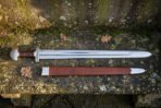 Hand forged high carbon steel Viking broadsword with round pommel, shown full length beside its brown leather scabbard on a stone ledge