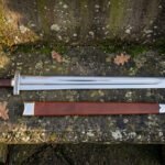 Hand forged high carbon steel Viking broadsword with round pommel, shown full length beside its brown leather scabbard on a stone ledge