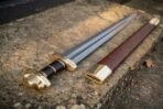 Hand forged high carbon steel Viking sword with brass lobed pommel and guard, angled on a stone step beside its leather scabbard