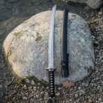 Hand forged high carbon steel wakizashi with black wrapped handle, standing upright against a large boulder beside its saya on gravel