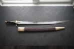 Hand forged Indian sword of talwar type with a mirror polished curved blade and brass disc pommel, laid on dark slate beside its brass mounted leather scabbard