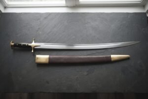 Hand forged Indian sword of talwar type with a mirror polished curved blade and brass disc pommel, laid on dark slate beside its brass mounted leather scabbard