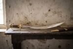 Hand forged Indian talwar with a deeply curved polished blade, rosewood grip and solid brass knuckle guard resting on a dark timber table against a pale plaster wall