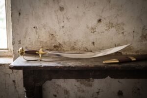 Hand forged Indian talwar with a deeply curved polished blade, rosewood grip and solid brass knuckle guard resting on a dark timber table against a pale plaster wall