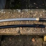Hand forged katana with a black cord wrapped tsuka and black lacquered saya laid along a lichen covered stone bench in soft low winter light
