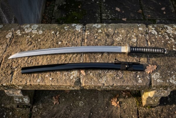 Hand forged katana with a black cord wrapped tsuka and black lacquered saya laid along a lichen covered stone bench in soft low winter light