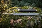 Hand forged langes messer with a polished straight backed blade and dark hardwood grip resting on a mossy boulder among oak leaves beside a brown leather sheath