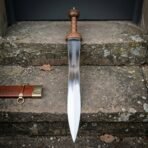 Hand forged Mainz gladius with a polished waisted leaf blade and ribbed cherry wood grip, standing upright on a lichened stone doorstep beside its tooled leather scabbard.