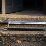 Hand forged medieval arming sword in polished high carbon steel with an etched Mjolnir panel, laid with its black scabbard on a lichened stone doorstep.