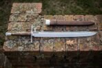 Hand forged messer sword with a polished straight backed blade and cream bone toned grip lying on a lichened red brick ledge beside a brown leather sheath