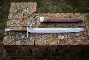 Hand forged messer sword with a polished straight backed blade and cream bone toned grip lying on a lichened red brick ledge beside a brown leather sheath