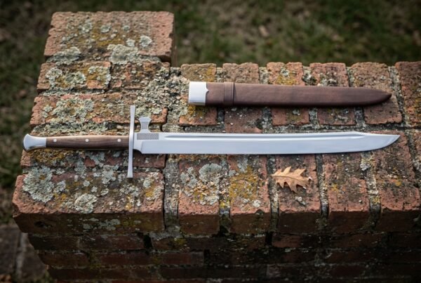 Hand forged messer sword with a polished straight backed blade and cream bone toned grip lying on a lichened red brick ledge beside a brown leather sheath