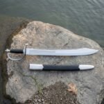 Hand forged naval cutlass with a broad polished steel blade, etched knotwork band, steel stirrup knuckle bow and ribbed black leather grip lying on a river smoothed granite rock beside its black scabbard