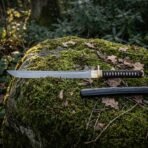 Hand forged ninja sword with a straight 1095 blade, angled tip and black cord wrapped grip lying on a moss covered woodland boulder beside its black saya