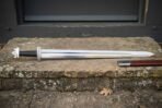 Hand forged Norse sword with broad polished blade, domed tea cosy pommel and brown leather scabbard lying on a worn stone doorstep in daylight