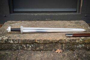 Hand forged Norse sword with broad polished blade, domed tea cosy pommel and brown leather scabbard lying on a worn stone doorstep in daylight