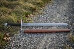 Hand forged Oakeshott type XII arming sword in polished high carbon steel with a wide fuller and black leather grip, lying on a gravel path beside its brown scabbard.