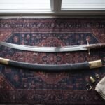 Hand forged Persian sword with a narrow crescent shamshir blade and walnut grip laid on a navy and rust Persian carpet beside its brass mounted leather scabbard