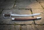 Hand forged pirate cutlass with a broad clipped point blade, fullered forte, steel knuckle bow and walnut slab grip laid on grey flagstone paving beside its brown leather scabbard