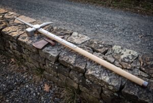 Hand forged polehammer with a polished steel hammer face, curved beak and top spike on a pale ash haft resting along a low drystone wall