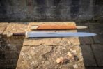 Hand forged ranger sword in high carbon steel with a leather wrapped grip and tan leather scabbard resting on a lichen covered stone slab outdoors
