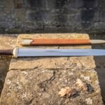 Hand forged ranger sword in high carbon steel with a leather wrapped grip and tan leather scabbard resting on a lichen covered stone slab outdoors