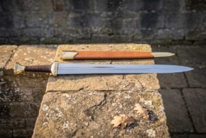 Hand forged ranger sword in high carbon steel with a leather wrapped grip and tan leather scabbard resting on a lichen covered stone slab outdoors