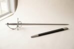 Hand forged rapier with a polished side ring guard and wire wrapped grip photographed on a pale studio backdrop beside its black leather scabbard