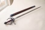 Hand forged renaissance rapier with an antiqued fullered blade and blackened swept hilt on a pale studio backdrop beside an oak leaf tooled leather scabbard