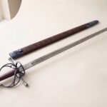 Hand forged renaissance rapier with an antiqued fullered blade and blackened swept hilt on a pale studio backdrop beside an oak leaf tooled leather scabbard