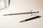 Hand forged renaissance short sword with a broad clipped point blade and polished knuckle bow hilt on a pale studio backdrop beside its black leather scabbard