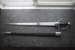Hand forged renaissance side sword with a shell plated ring guard and pinned ebony slab grip laid on dark slate beside its brown leather scabbard