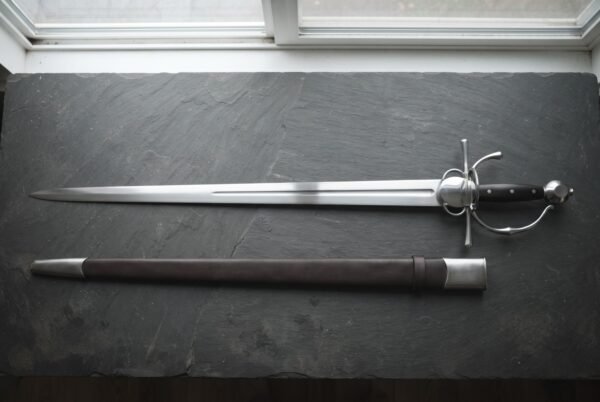 Hand forged renaissance side sword with a shell plated ring guard and pinned ebony slab grip laid on dark slate beside its brown leather scabbard