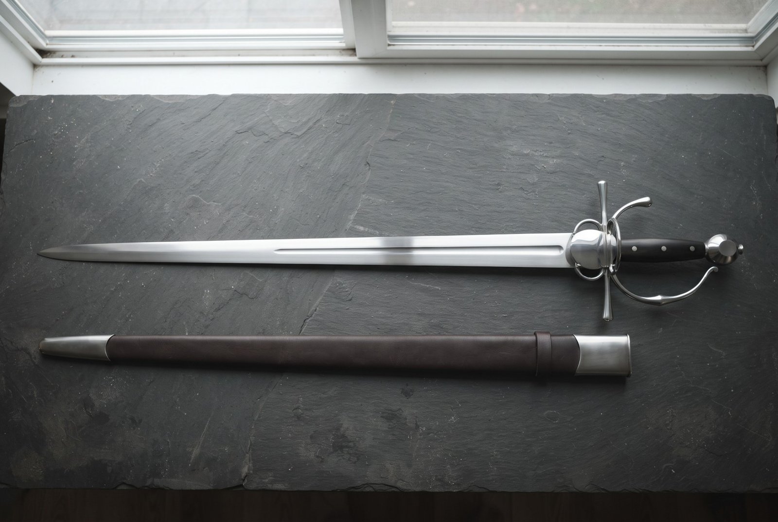 Hand forged renaissance side sword with a shell plated ring guard and pinned ebony slab grip laid on dark slate beside i Hand forged renaissance side sword with a shell plated ring guard and pinned ebony slab grip laid on dark slate beside its brown leather scabbard