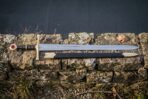 Hand forged runic sword with a darkened Elder Futhark blade inscription, deep red leather grip and steel disc pommel lying on a dark drystone wall