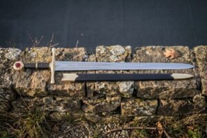 Hand forged runic sword with a darkened Elder Futhark blade inscription, deep red leather grip and steel disc pommel lying on a dark drystone wall