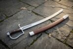 Hand forged sabre sword with a broad polished curved blade, steel knuckle bow hilt and rosewood slab grip resting on grey flagstone paving beside its brown leather scabbard