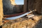 Hand forged scimitar sword with a mirror polished curved blade and brass hilt resting on a sunlit stone ledge beside its tooled leather scabbard and a whetstone