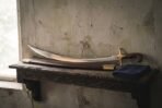 Hand forged shamshir sword with a narrow curved polished blade and one piece rosewood grip resting on a carved timber shelf beside its brass mounted leather scabbard