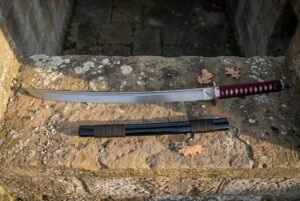 Hand forged short samurai sword with a deep red cord wrapped grip on a weathered stone step, its black saya with brown cord wrap laid below the polished blade