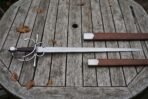 Hand forged side sword with a broad fullered blade and polished swept hilt lying on a weathered timber garden table beside its brown leather scabbard