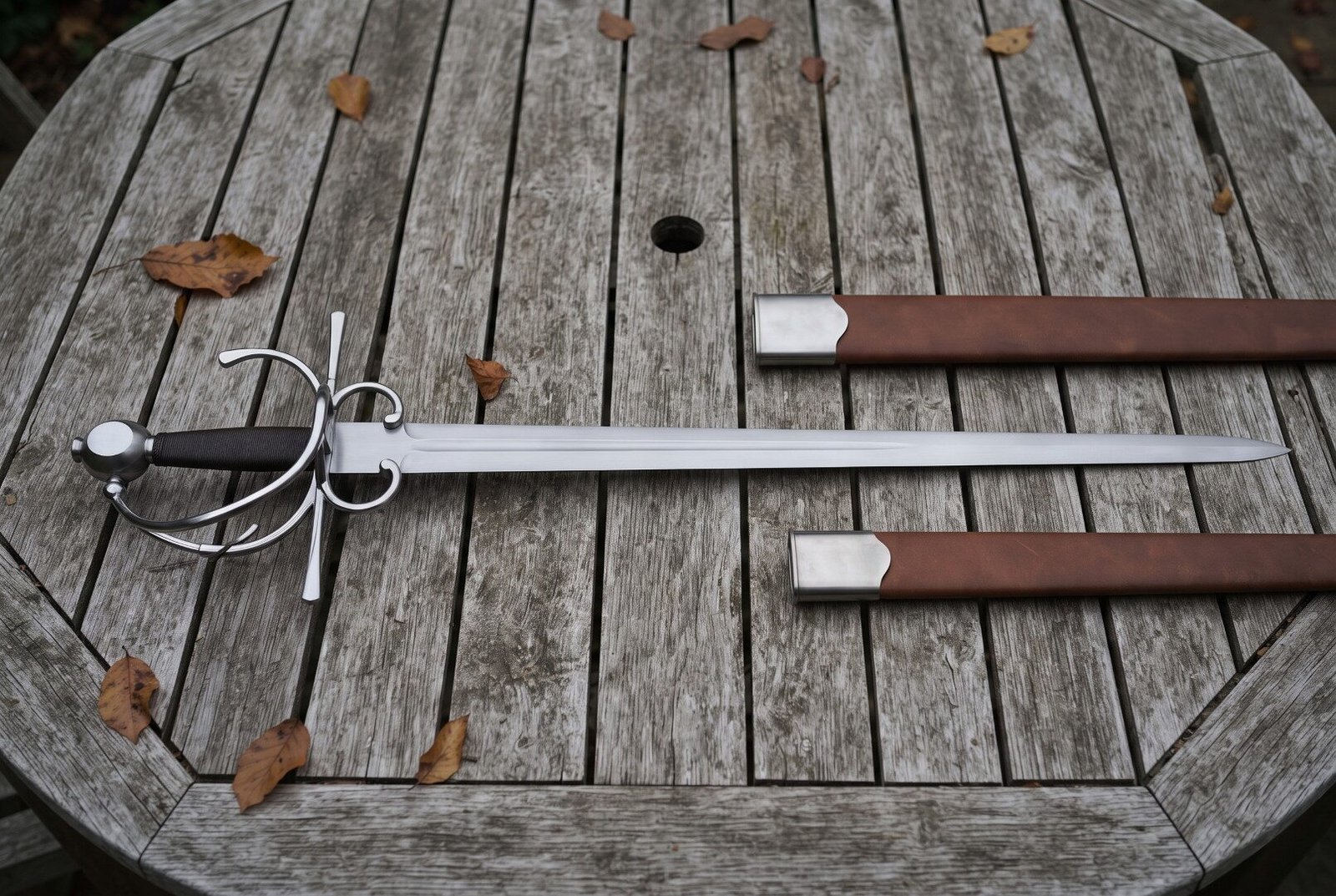 Hand forged side sword with a broad fullered blade and polished swept hilt lying on a weathered timber garden table besi Hand forged side sword with a broad fullered blade and polished swept hilt lying on a weathered timber garden table beside its brown leather scabbard